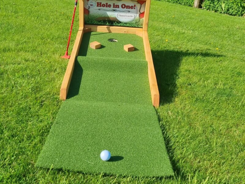 This is a picture of our new Hole in One Game. It is made of an artificial grass panel and wood. It is bright, being red and white. The hole is in the style of crazy golf. This game is for hire for your event.