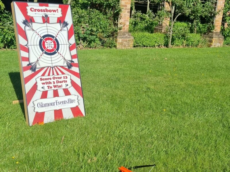 One of our games is shown which is the 'Crossbow' game. It is a red and white striped board on an angle. There is a target on the front. There is a crossbow that you can use to try and hit the target with arrows. This game is for hire.